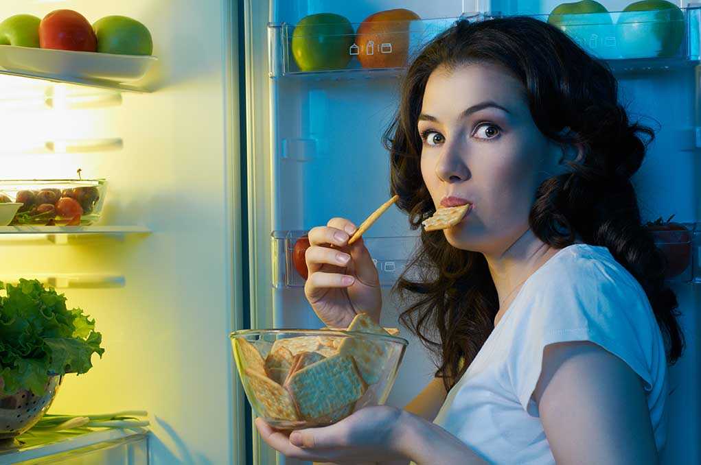 shutterstock_111599705.jpg A woman eating a snack while looking at the camera, standing in front of an open refrigerator