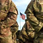 Soldiers in camouflage stand with hands behind backs facing a U.S. flag