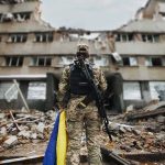 Soldier with rifle stands before a destroyed building holding a blue and yellow flag