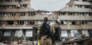 Fighting Intensifies In Kostiantynivka Soldier with rifle stands before a destroyed building holding a blue and yellow flag