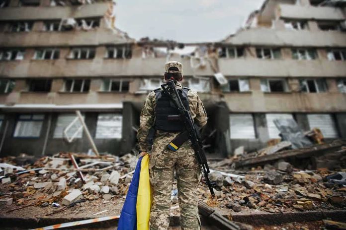 Soldier with rifle stands before a destroyed building holding a blue and yellow flag