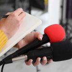 SHOCKING ‘Birth Tourism’ Scheme Exposed in Texas Journalist holding two microphones while writing in a notebook