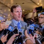 Man in suit speaks to reporters surrounded by microphones